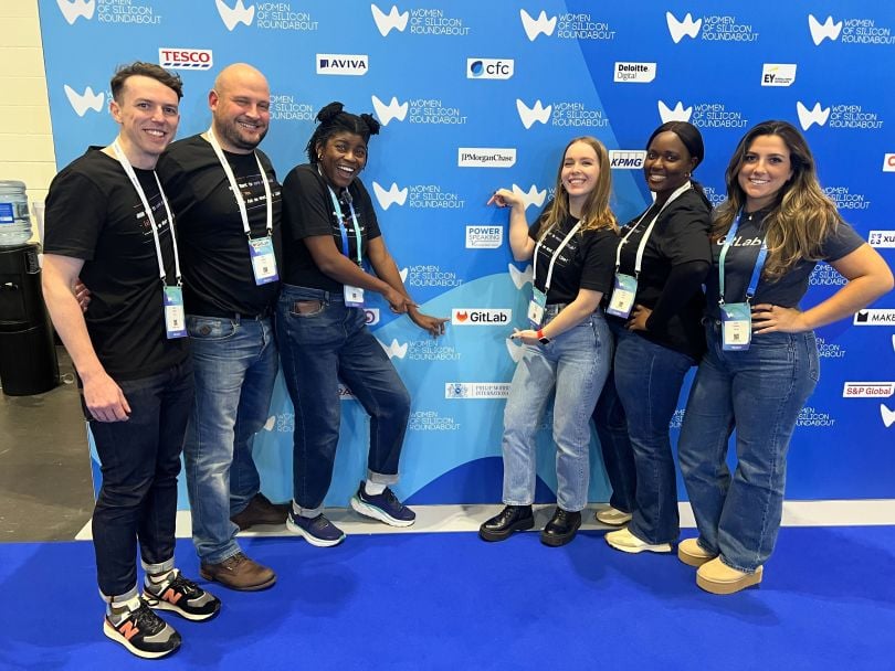 Group photo of GitLab team members in front of a step-and-repeat backdrop at a GitLab-sponsored event, pointing to the GitLab logo on the backdrop. 