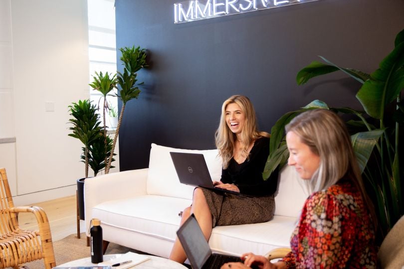 Photo of two coworkers sitting on couches holding laptops; one woman is laughing.