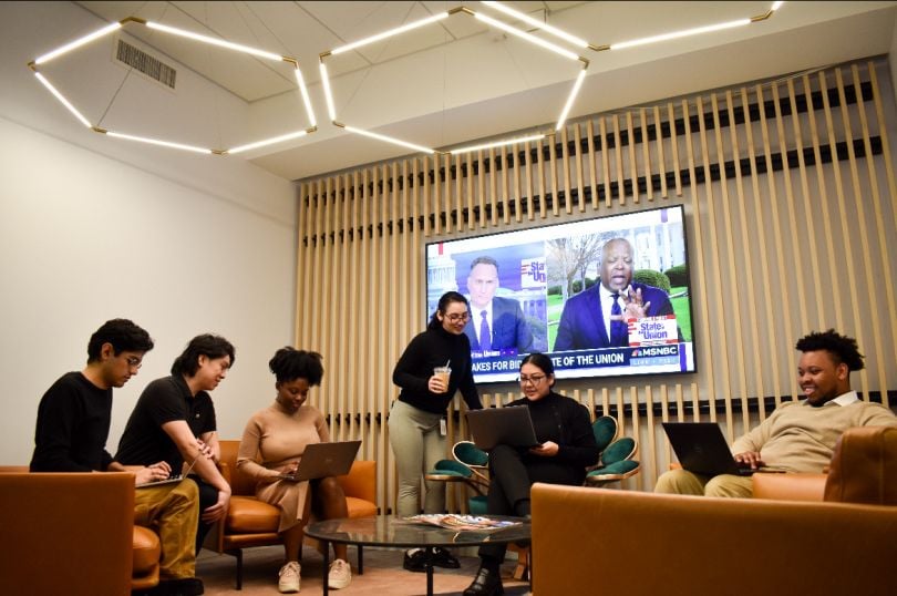 NBCUniversal team members in a meeting sitting on couches and lounge chairs, with MSNBC channel on a TV on the wall. 