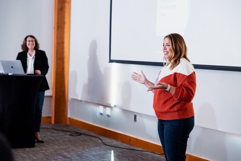 A leader stands at the front of a room, smiling and addressing a team, while Jennifer Guisbert stands, smiling attentively, running the presentation on a laptop at the lectern nearby. 