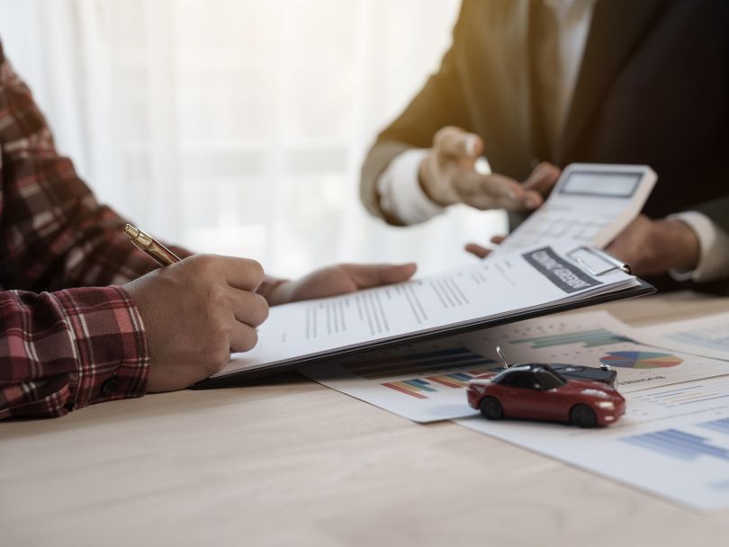  A customer signs the contract to purchase a new car as the salesperson gestures to a number on a calculator.