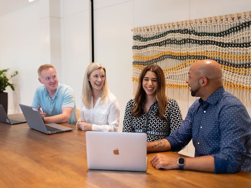 Telesign teammates sit next to each other at a table at the company’s 2024 Revenue Kickoff event, looking at laptops together and smiling. 