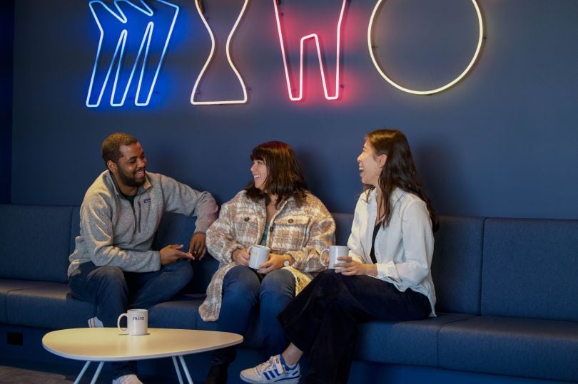 Candid photo of Miro team members sitting on a upholstered bench, talking with fluorescent Miro logo on wall behind them.