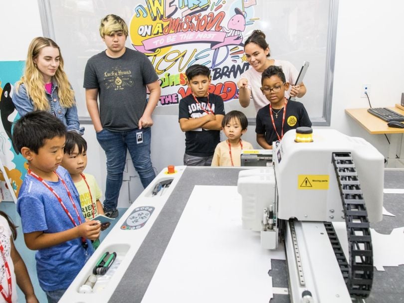 Munchkin employees show a group of kids a high-tech printer in their office. The wall mural in the background reads, “We are on a mission to be the most loved lifestyle brand in the world.”