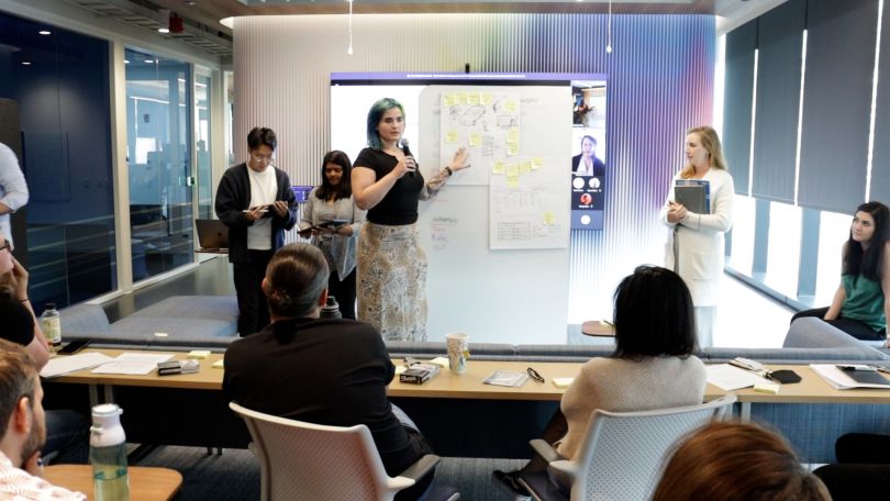 Woman with microphone presenting next to board with sticky notes in front of group of seated colleagues