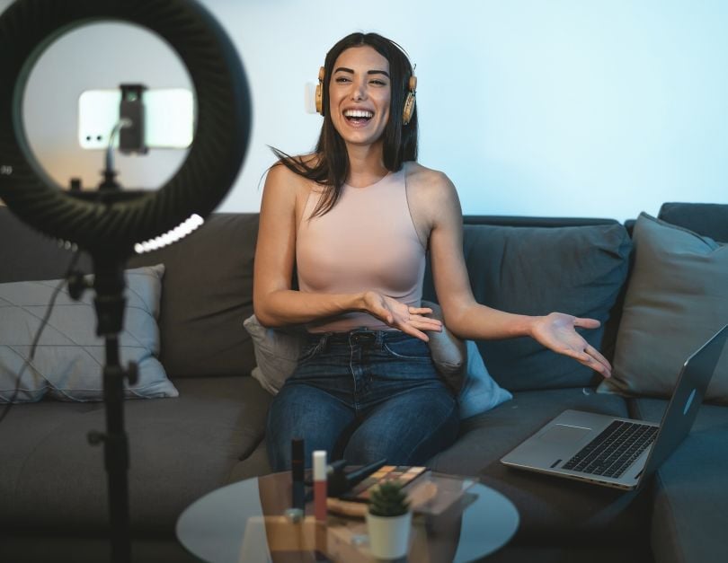 An influencer smiles and gestures at the laptop next to her while looking into a cell phone mounted on a standing ring light.