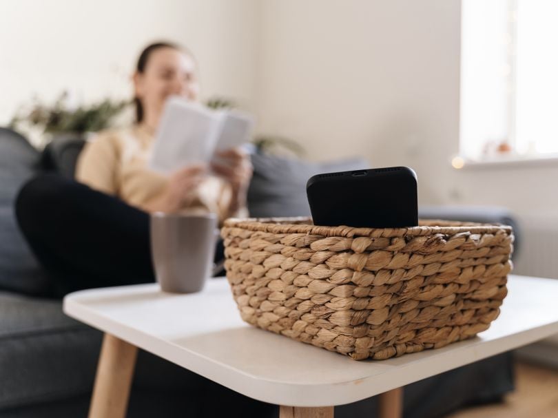 A woman leaves her cell phone in a basket while she reads on the couch.