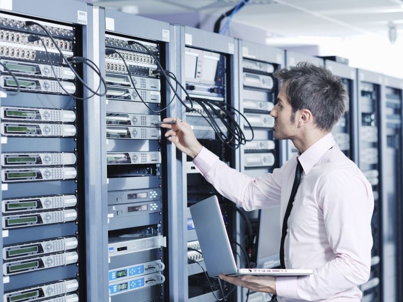 An engineer inspects a bank of servers while holding a laptop.