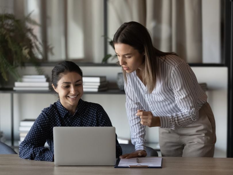 An employee smiles while receiving feedback from a colleague as they look at her laptop together.