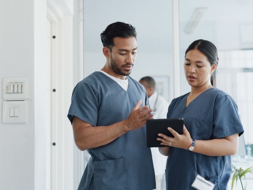 Two healthcare workers discuss something while referencing a tablet in front of them.