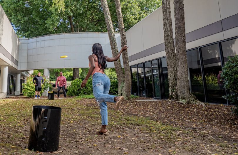 A woman in the foreground with her back to the camera throws a frisbee across a courtyard toward two men. The group is playing Kanjam.