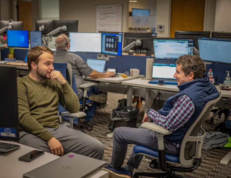 Two men sit in desk chairs and face each other to talk