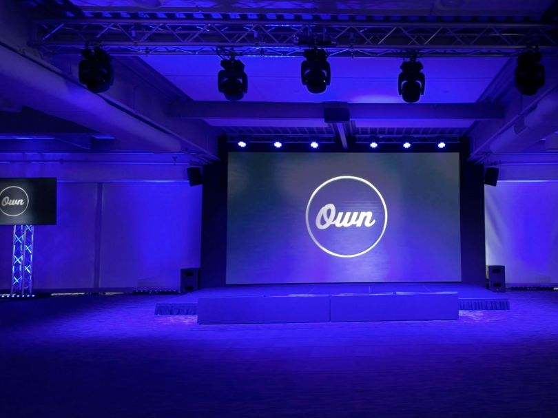 A photo of Own Company’s Radio City meeting auditorium, featuring a stage with a large screen and flanking televisions that display the company logo.