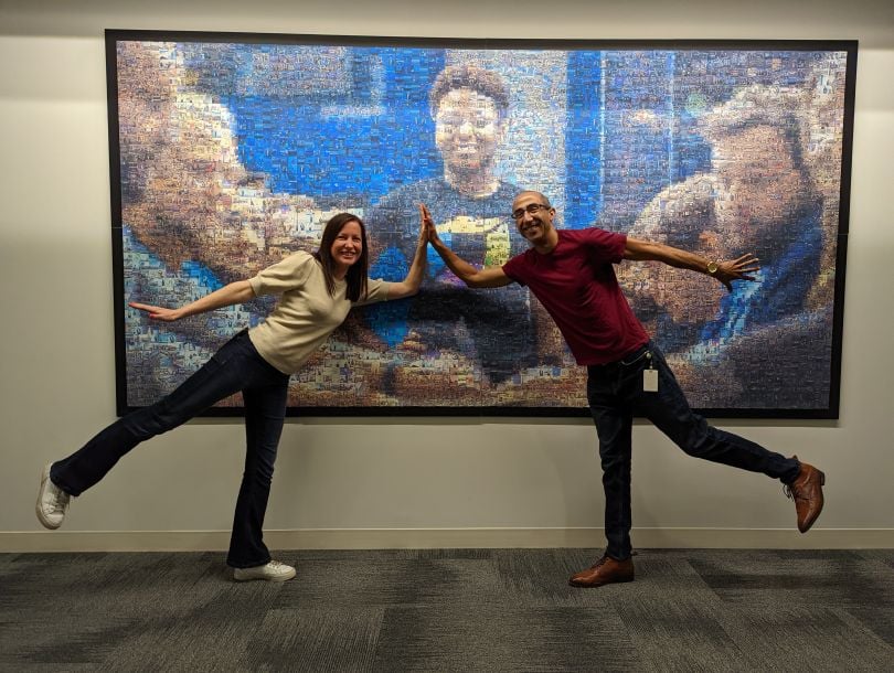 John Rocker and a colleague pose in a high-five position (each leaning forward on one foot) in front of a large wall-hung photo. 