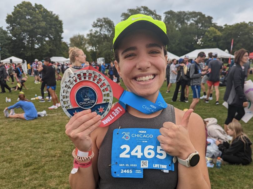 Photo of Raquel Zanoni after completing a Chicago race, wearing a bib and holding a medal, giving a thumbs-up sign.