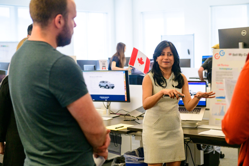 CarGurus team members listening as colleague explains, standing near open-seating desks.