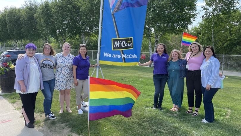 McCain Foods employees stand outside in the grass next to an LGBTQIA+ pride flag and a banner that says “McCain: We are McCain.”