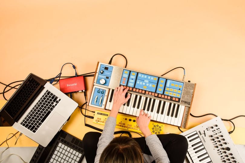 Overhead view of person sitting cross-legged on the floor playing a keyboard and surrounded by a laptop and other sound equipment. 