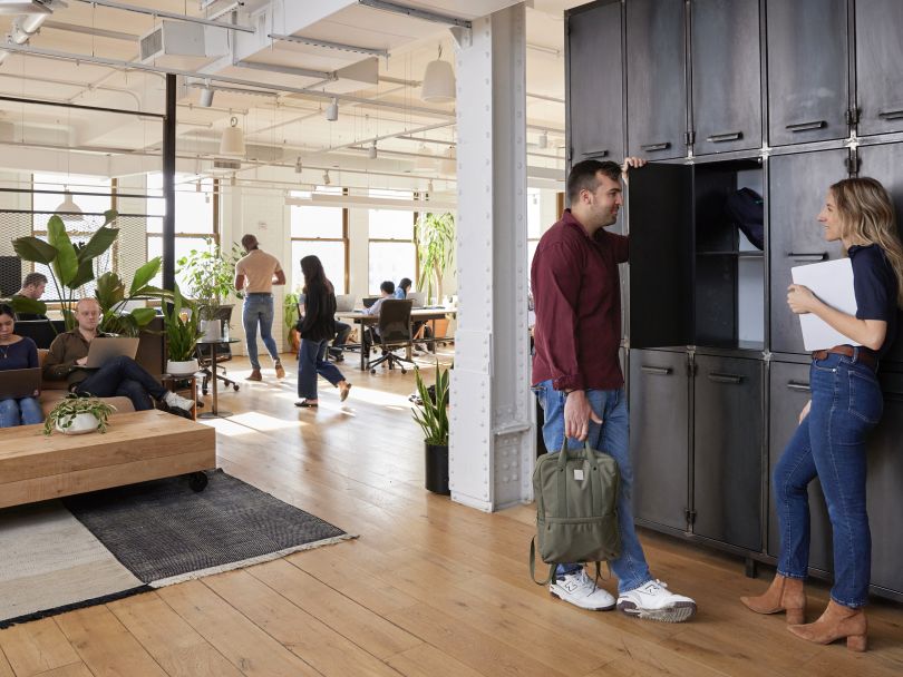 Riskified employees chat at a bank of lockers in the office as other employees work in the background.