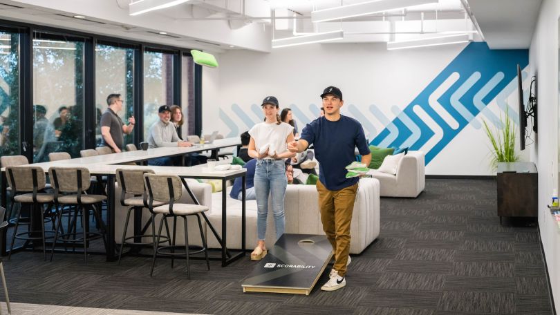 Scorability team members in the office, playing a game of cornhole near a gathering area.