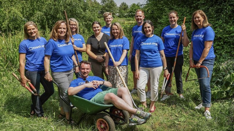 A group of Toasters in Ireland wearing company-branded T-shirts stand with gardening tools in a field, ready for a volunteer event with the company’s philanthropic and social impact branch, Toast.org. 