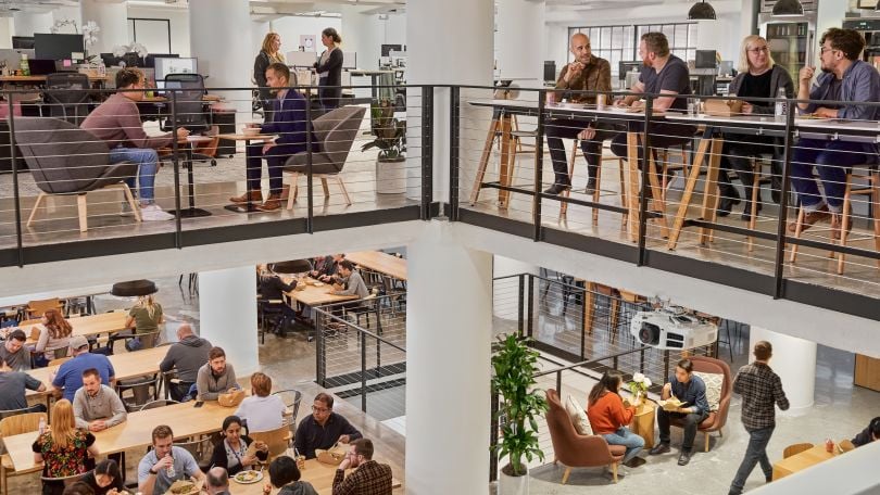 A photo of people working in an open office space or enjoying lunch. The view shows two floors through an open atrium. 