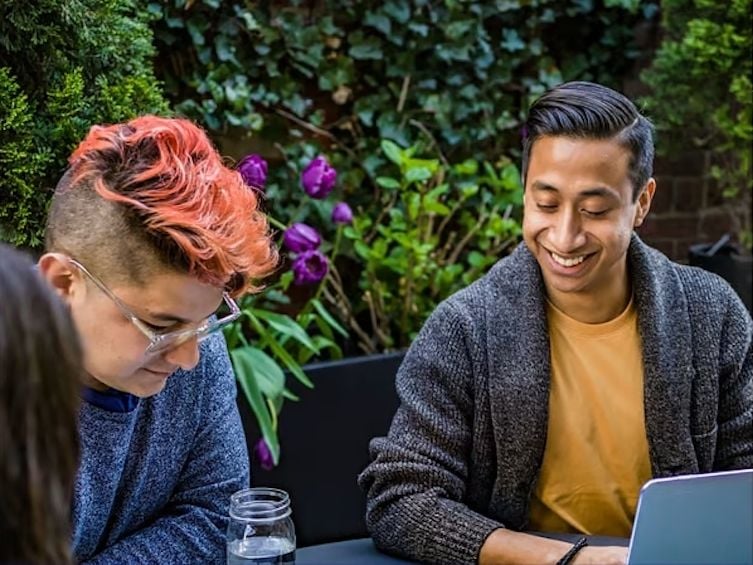 Two people sitting at a table with a laptop. Behind them, there is a large shrub and some flowers.