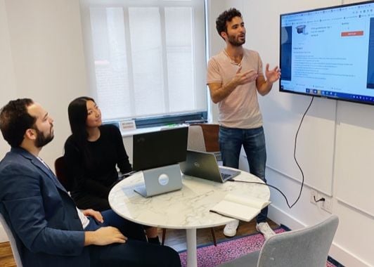 One of Torch's employees gesturing to a TV screen displaying some text. Two other people are sitting at a table with a laptop and watching him speak.