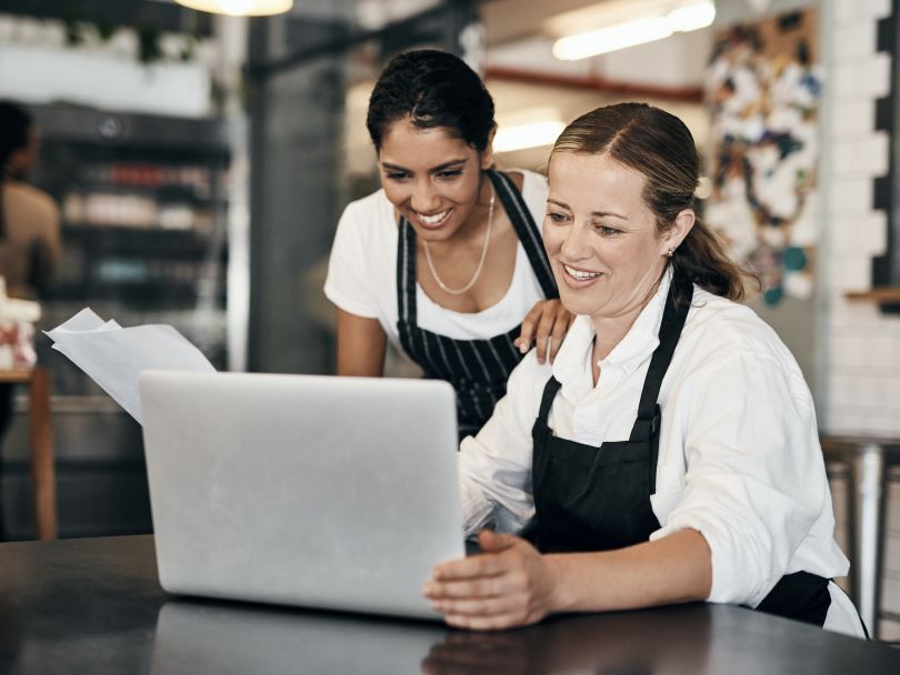 Two restaurant employees smile as they look between some papers and a laptop.