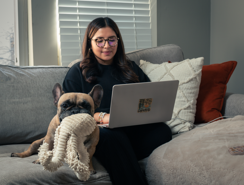 A remote VelocityEHS employee sits on couch with laptop, a dog laying next to her holding a toy in its mouth.