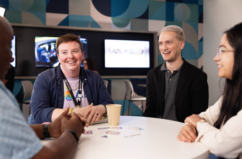 A group of Alliant employees sit around a table, smiling and talking. One person wears a PRIDE badge and various stickers lay on the table.