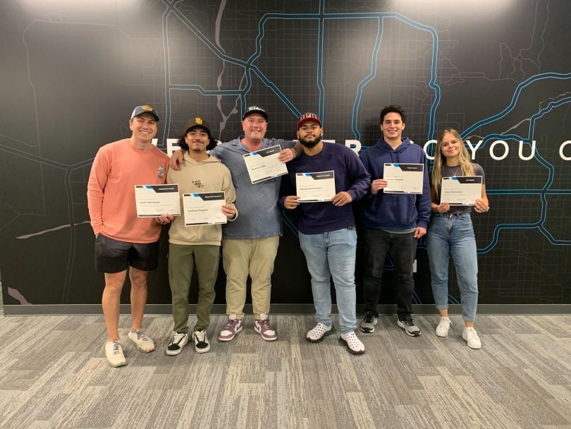 Six Arrive employees stand in front of a wall featuring a map-style graphic. Each person holds a certificate.