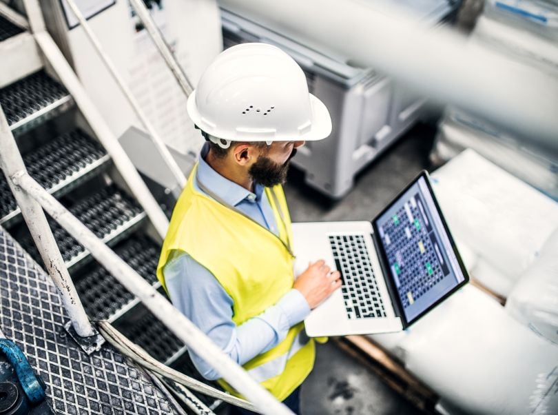 A technician wearing a hardhat and vest and standing amidst machinery checks his laptop to see how machines are performing.