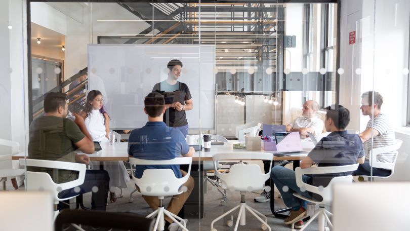 monday.com team members sitting around a conference table in a glass-walled meeting room. 