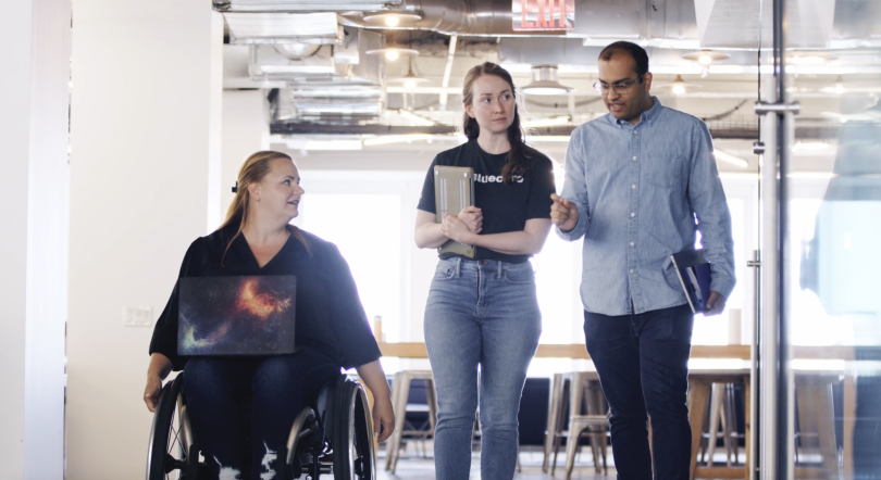 Three of Bluecore's employees walking down a hall and talking. From left to right: A woman in a wheelchair, a woman holding a clipboard and a man.