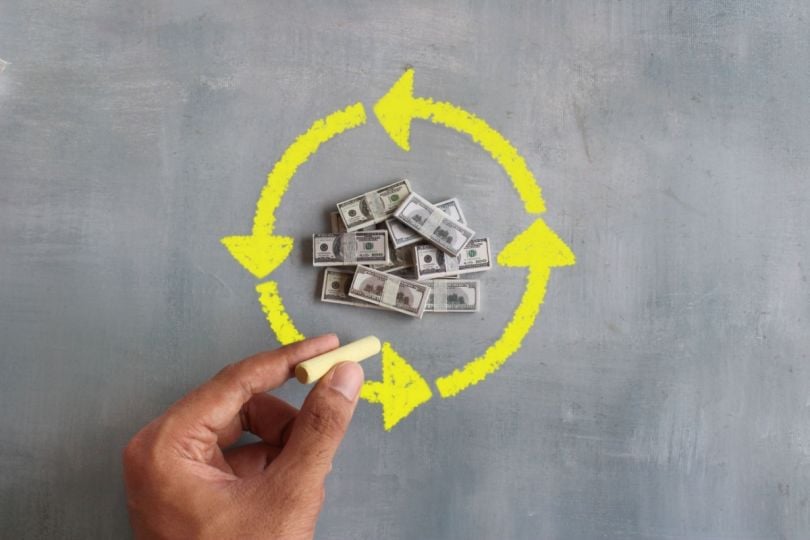 A hand drawing a circular arrow around miniature dollar bills.