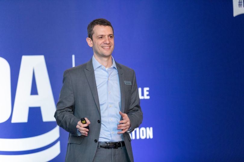 Ben Flusberg stands on stage at a conference in front of a blue backdrop with obscured white lettering, presenting and smiling.