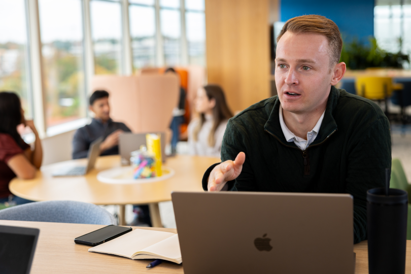 Cox Automotive employee sits behind a laptop and speaks to someone out of the photo, with colleagues chatting in the background in a communal seating area.