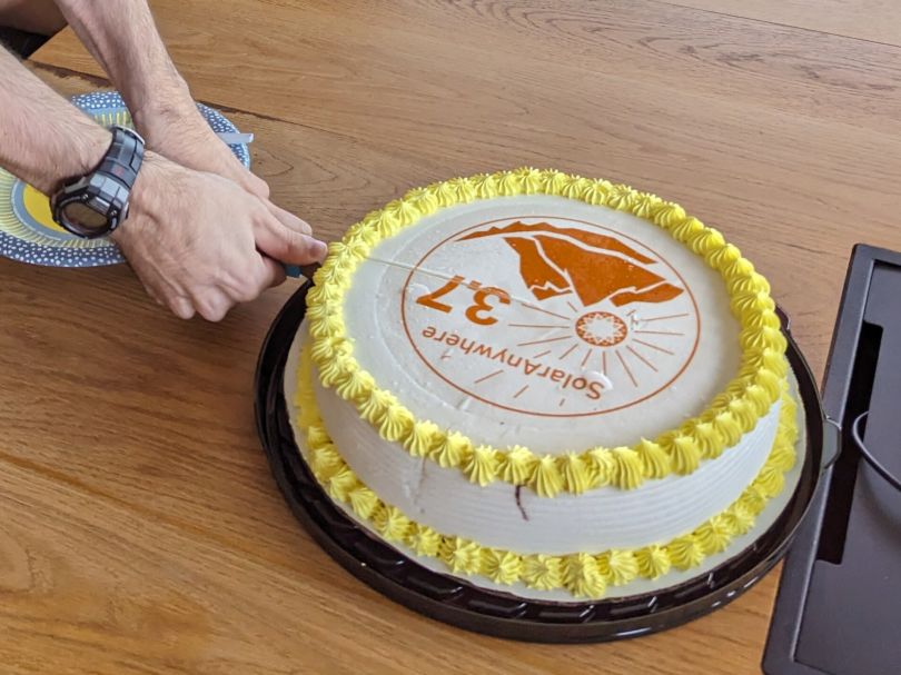 A cake labeled SolarAnywhere 3.7 with a mountain logo and a sunburst is cut by a person wearing a watch and holding a knife with both hands.