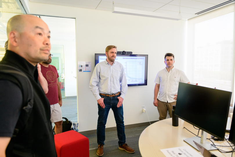  Photo of Jason Prentice and team members holding a standing meeting in a room with data on a screen on the wall and computers in the center.