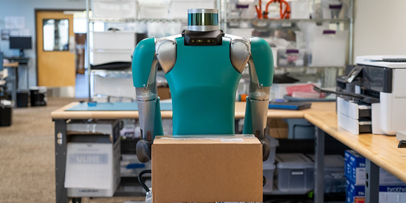 The humanoid robot, Digit, carrying a cardboard box in an office setting.
