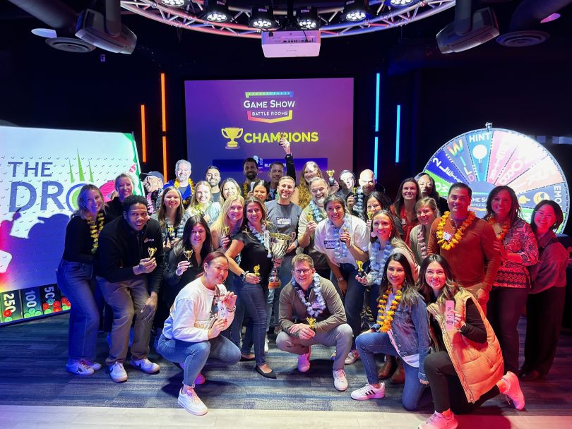 A group of more than 30 Ellevation employees pose in front of large screens, one of which reads “Game Show Battle Room Champions,” and a game show wheel.