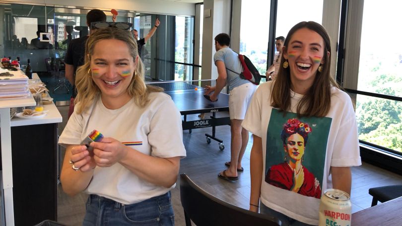 A photo of two people in white t-shirts, one with Frida Kahlo on the front, stand next to one another laughing. They both have small rainbows on their cheeks for a company LGBTQ Pride event. 