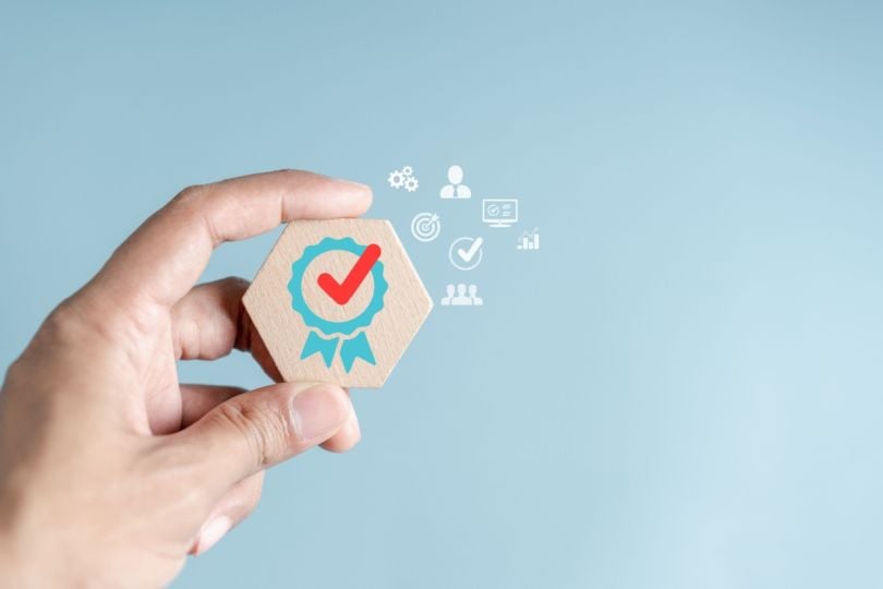 A hand holding a wooden block with a quality assurance badge symbol and various business icons floating above it.