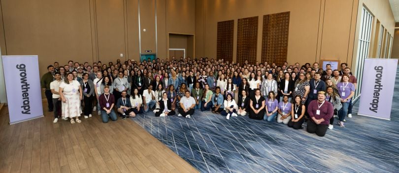 The Grow Therapy team poses in a ballroom or lobby flanked by two standing signs with the company’s name printed on them.