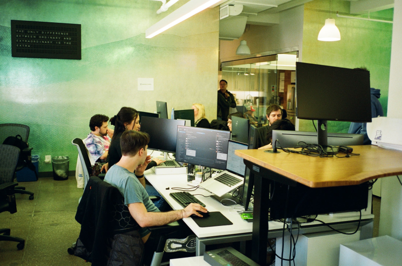 Photo of Pinwheel team members at bench desk seating working on computers side by side and across from colleagues