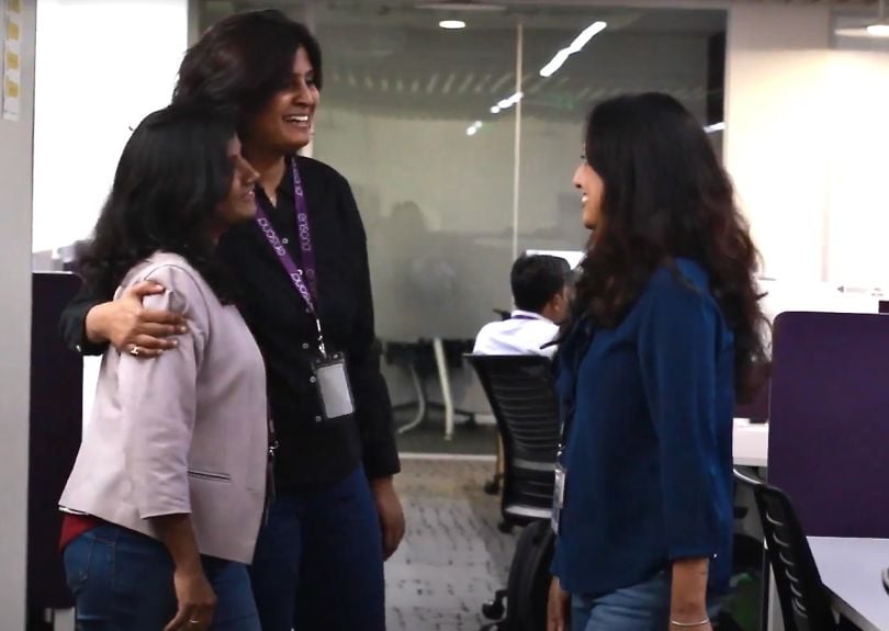 Candid photo of two women team members smiling and side-hugging as they talk to another colleague