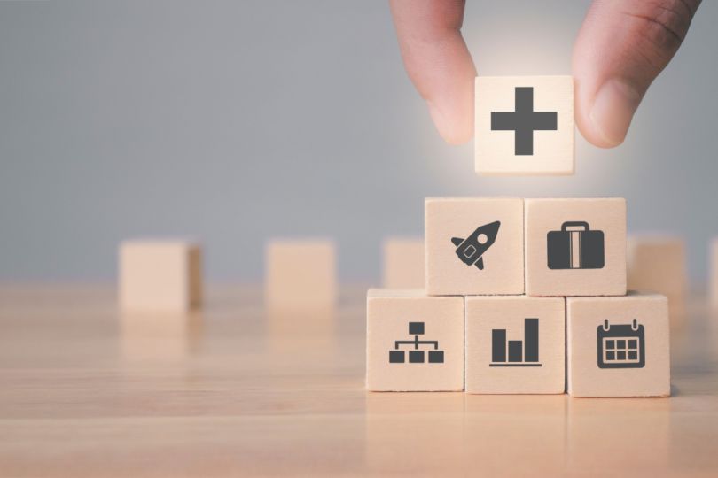 A person placing a wooden block with a plus sign on it on top of other blocks displaying various industry and business icons.