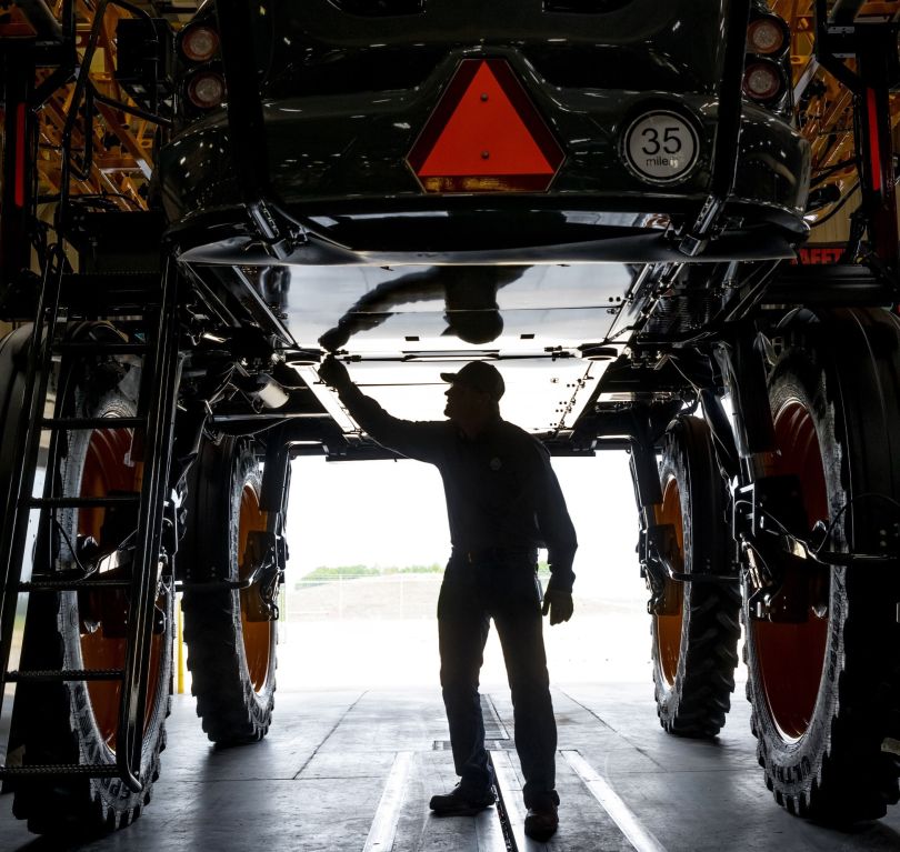 John Deere team member stands under large equipment, inspecting its underbelly. 