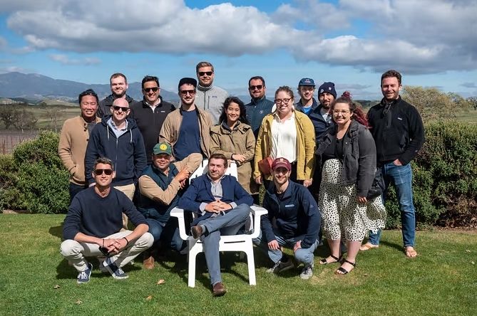 Team of 16 people in outdoor setting with green grass and partially cloudy sky